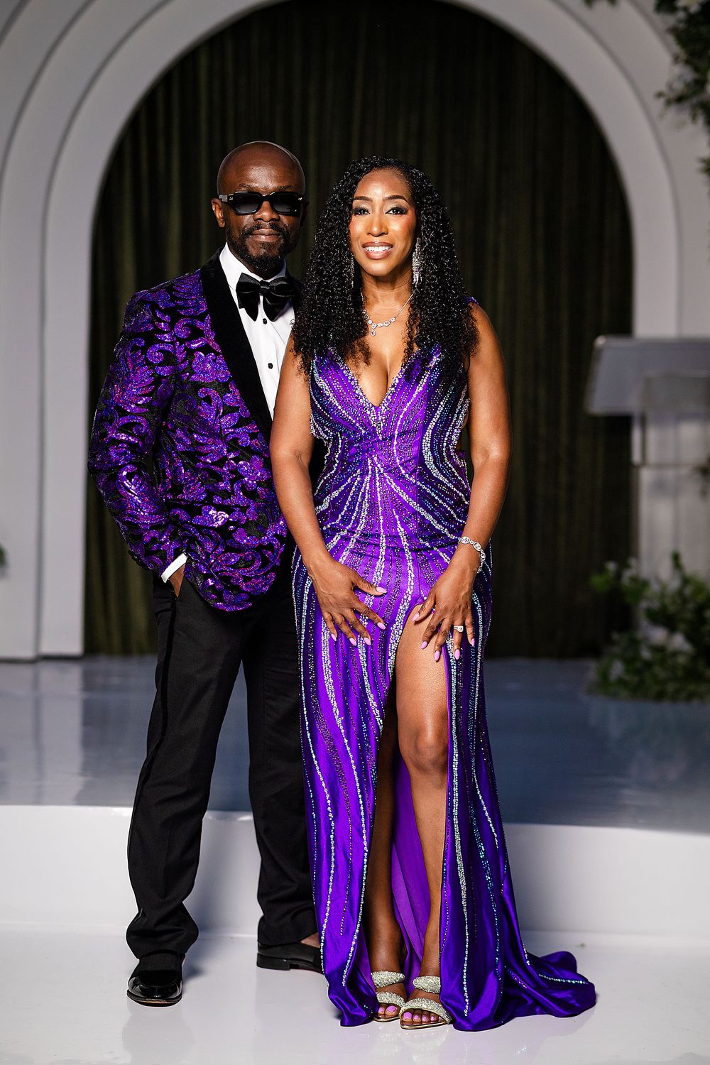 A couple in matching purple outfits, man in floral blazer and woman in beaded gown with slit, posing elegantly.