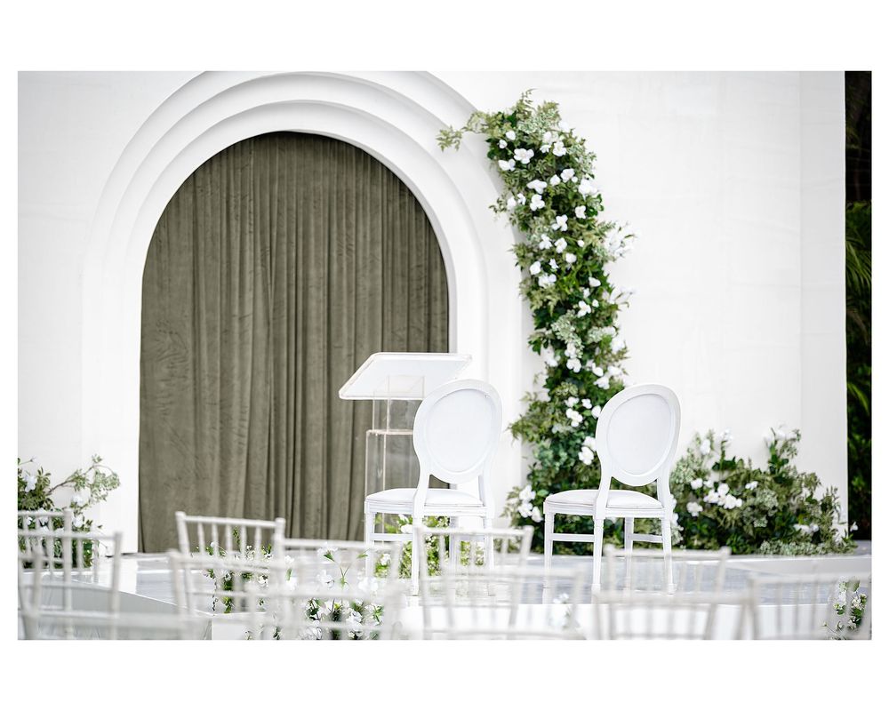 Elegant outdoor wedding ceremony setup with white chairs, floral arch, and green curtain backdrop.