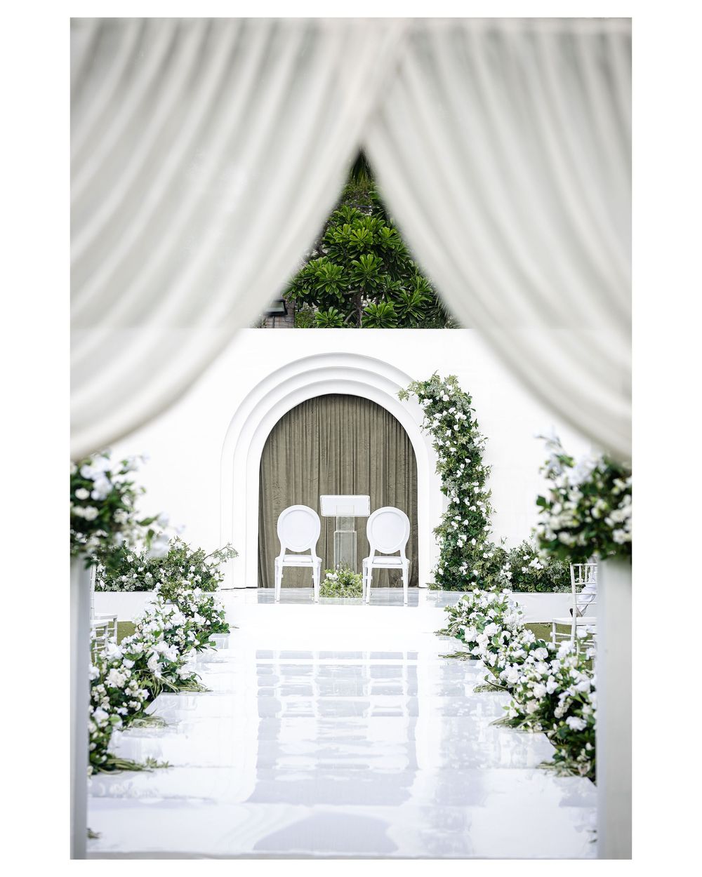 An elegant all-white outdoor wedding ceremony setup with floral arrangements, white chairs, and an arched backdrop framed by drapes.
