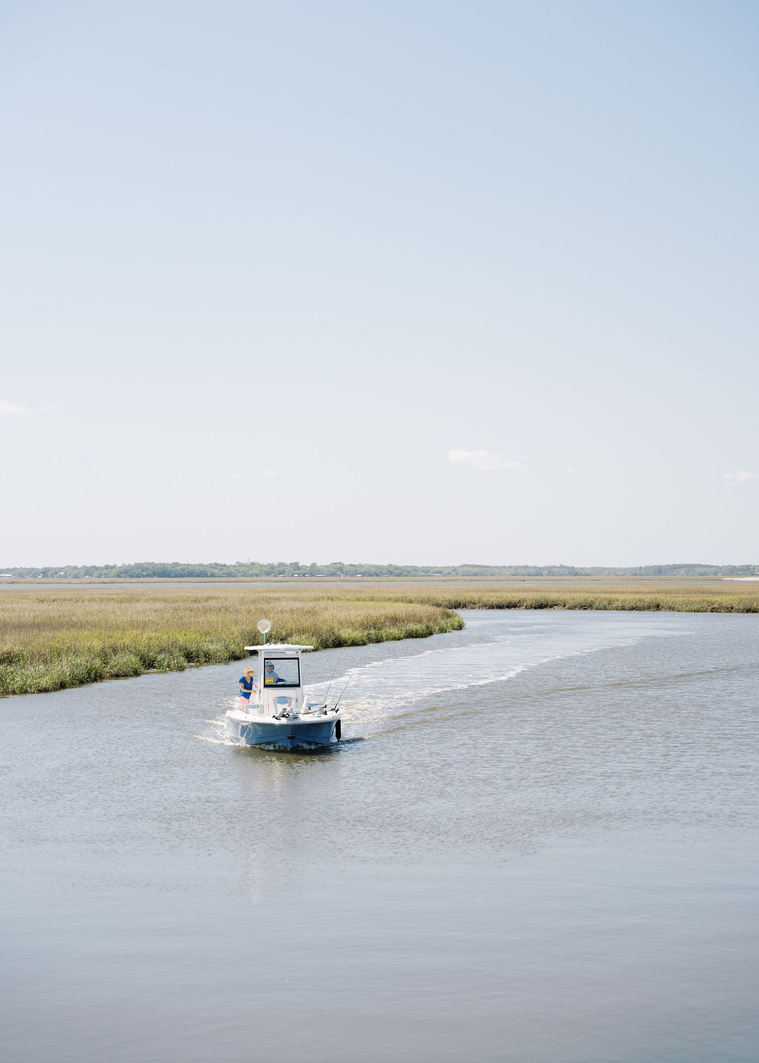Small boat cruising through a calm coastal marsh waterway under a clear blue sky.
