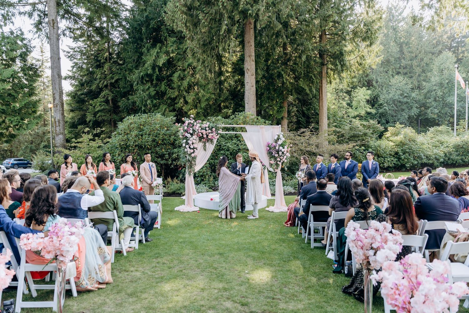 Outdoor garden wedding ceremony with pink floral decorations and guests seated on white chairs.