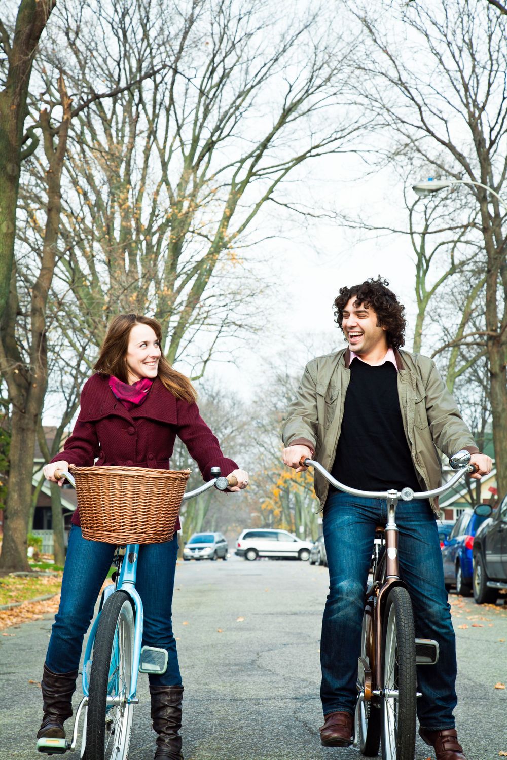 A couple rides bicycles together on an autumn street lined with bare trees.during their engagement session