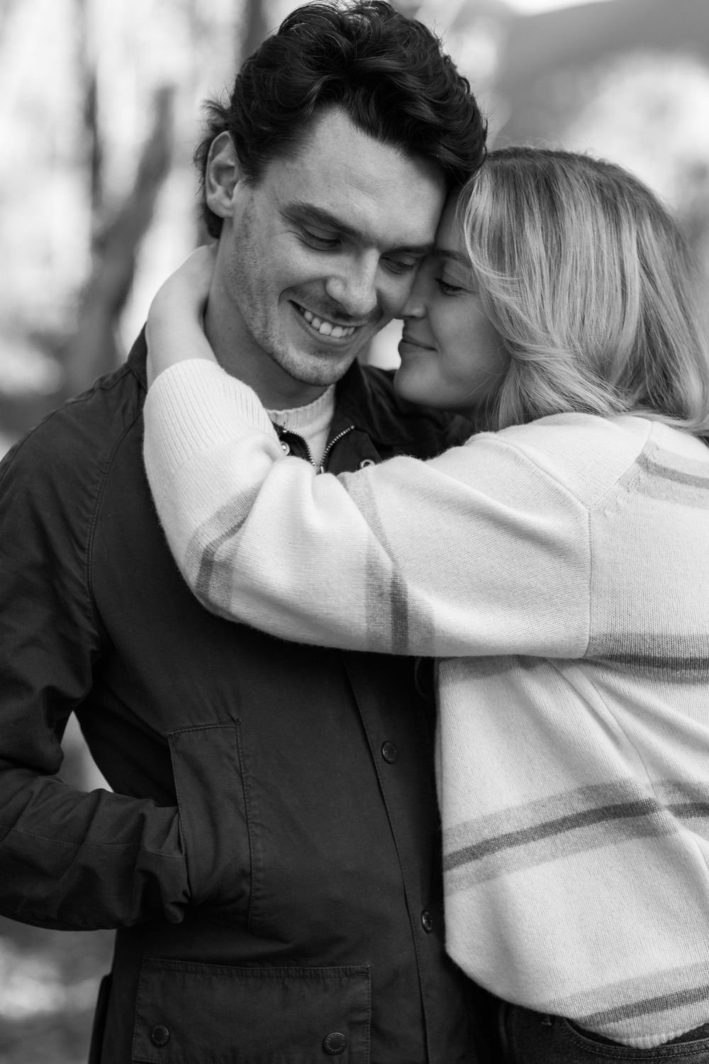 Black and white photo of a smiling man being embraced and kissed on cheek by a blonde woman outdoors.