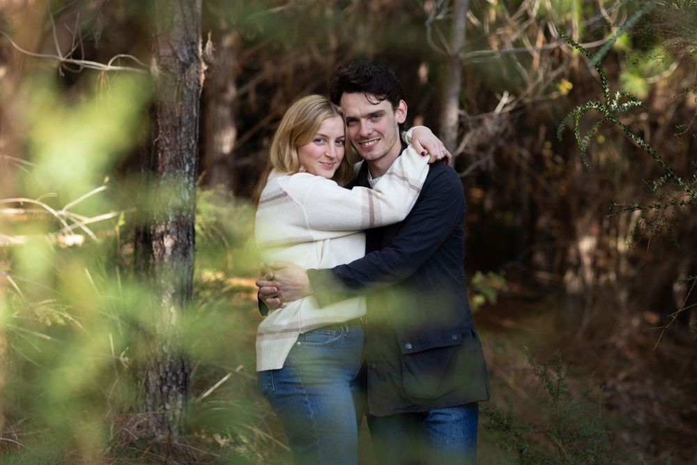 Couple embracing and smiling in a forest setting surrounded by green trees and autumn foliage.