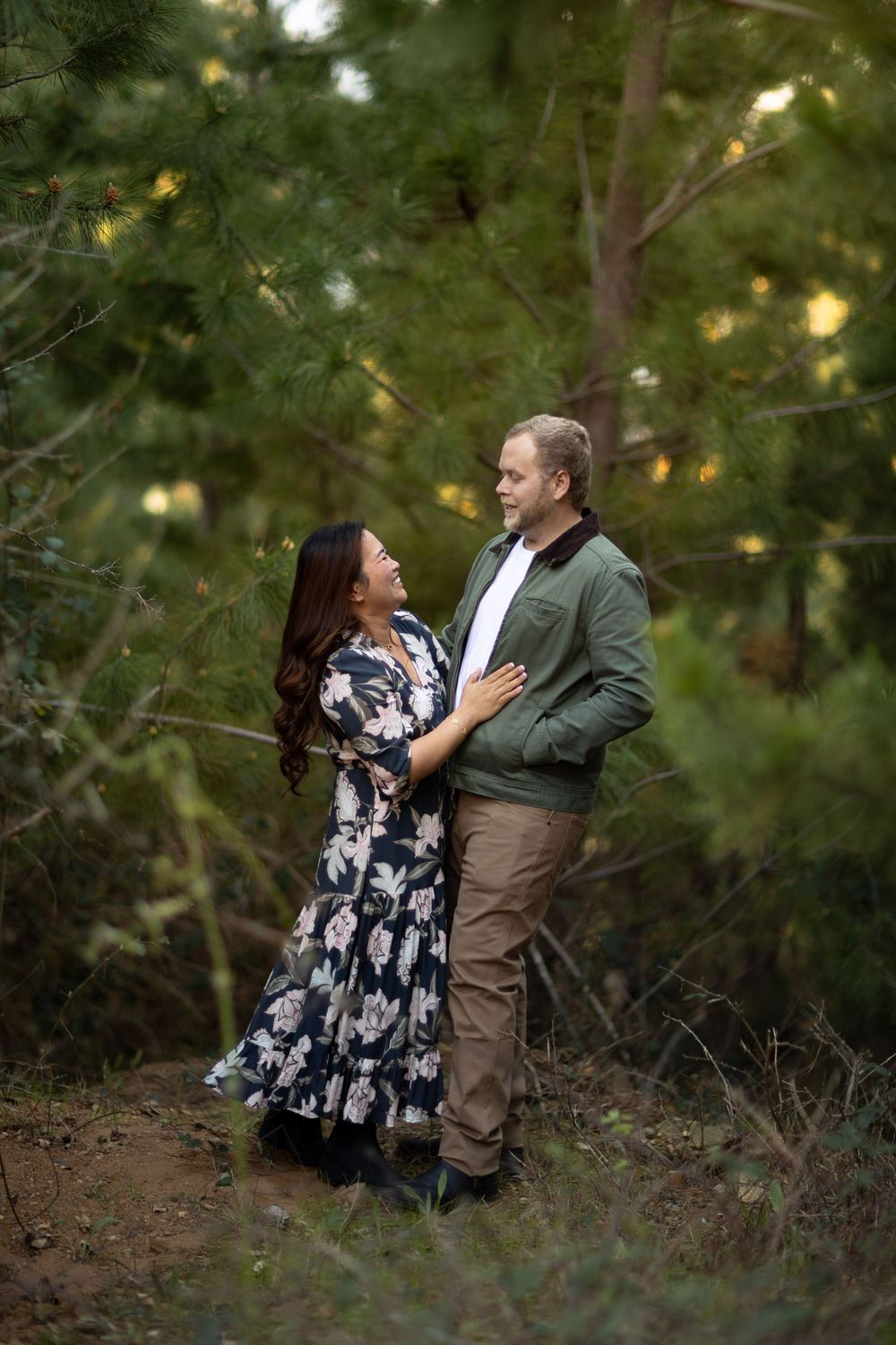 Couple embracing and gazing at each other in a lush green forest setting, woman in floral dress, man in green jacket.