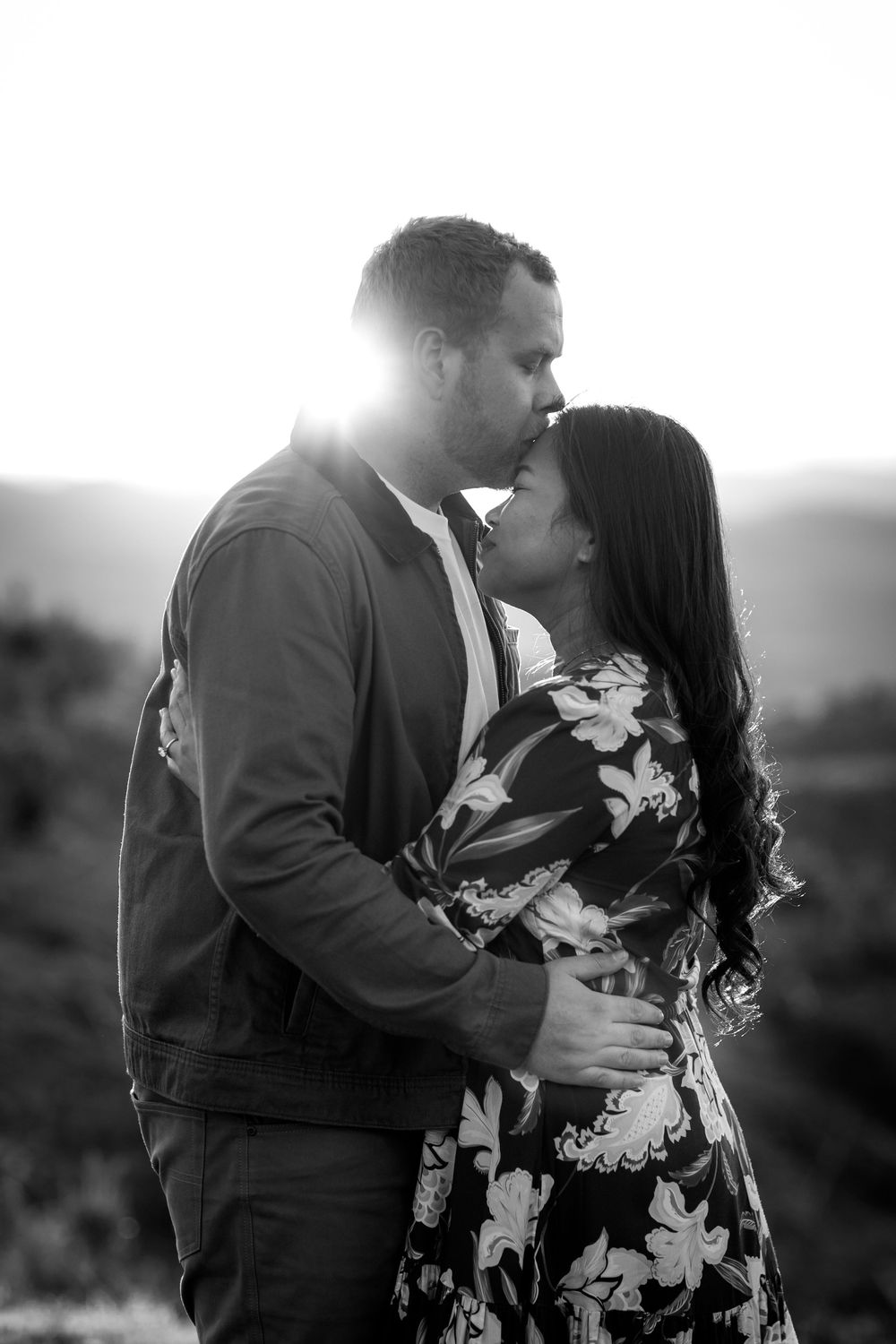 Black and white photo of a couple sharing a tender kiss outdoors with sunlight glowing behind them.