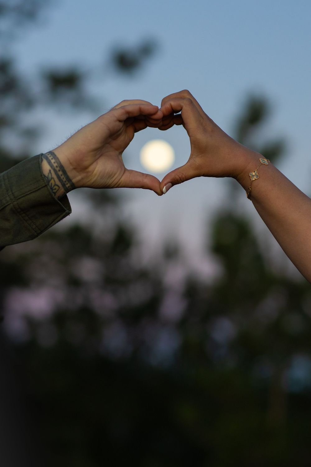 Two hands forming a heart shape with a full moon glowing in the center against a twilight sky.