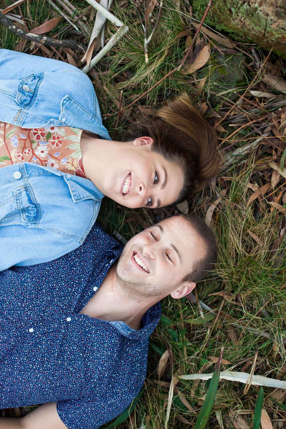 A couple lying in the grass wearing casual denim clothes share genuine smiles on a sunny day during their engagement session.