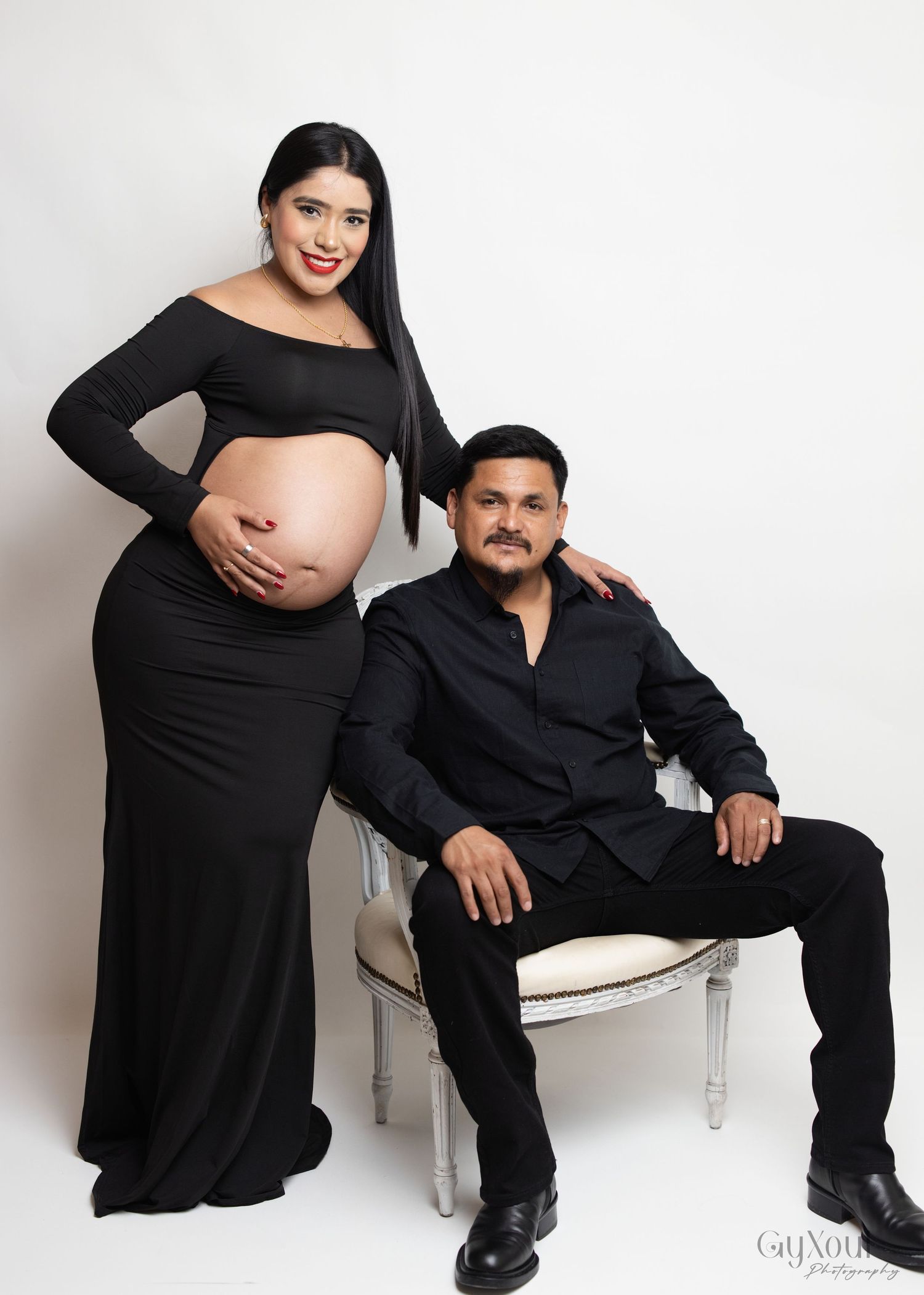 A maternity photoshoot featuring a couple in black attire against a white backdrop with an elegant chair.