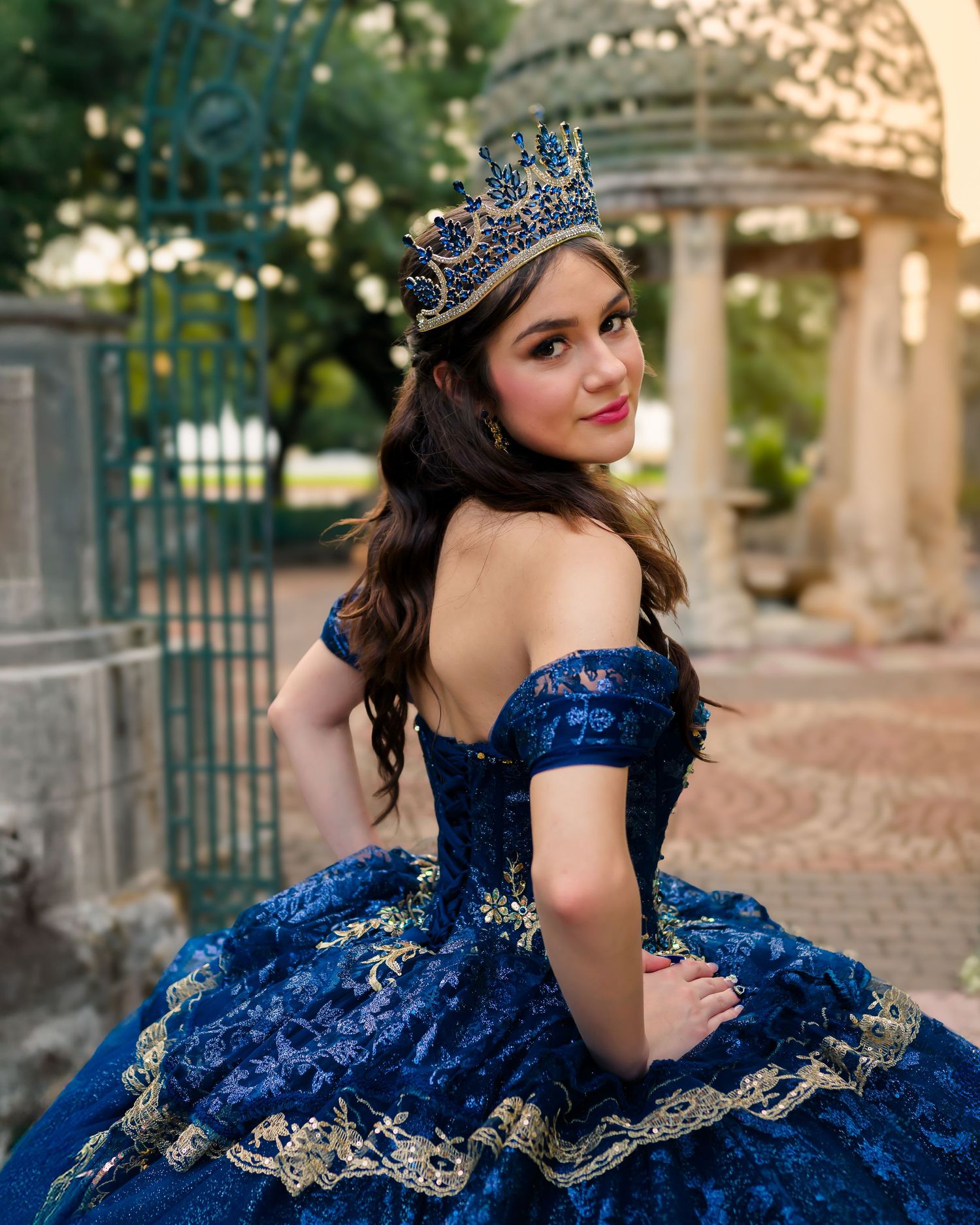 A glamorous royal blue quinceañera dress with gold embellishments sparkles in an outdoor photoshoot session.