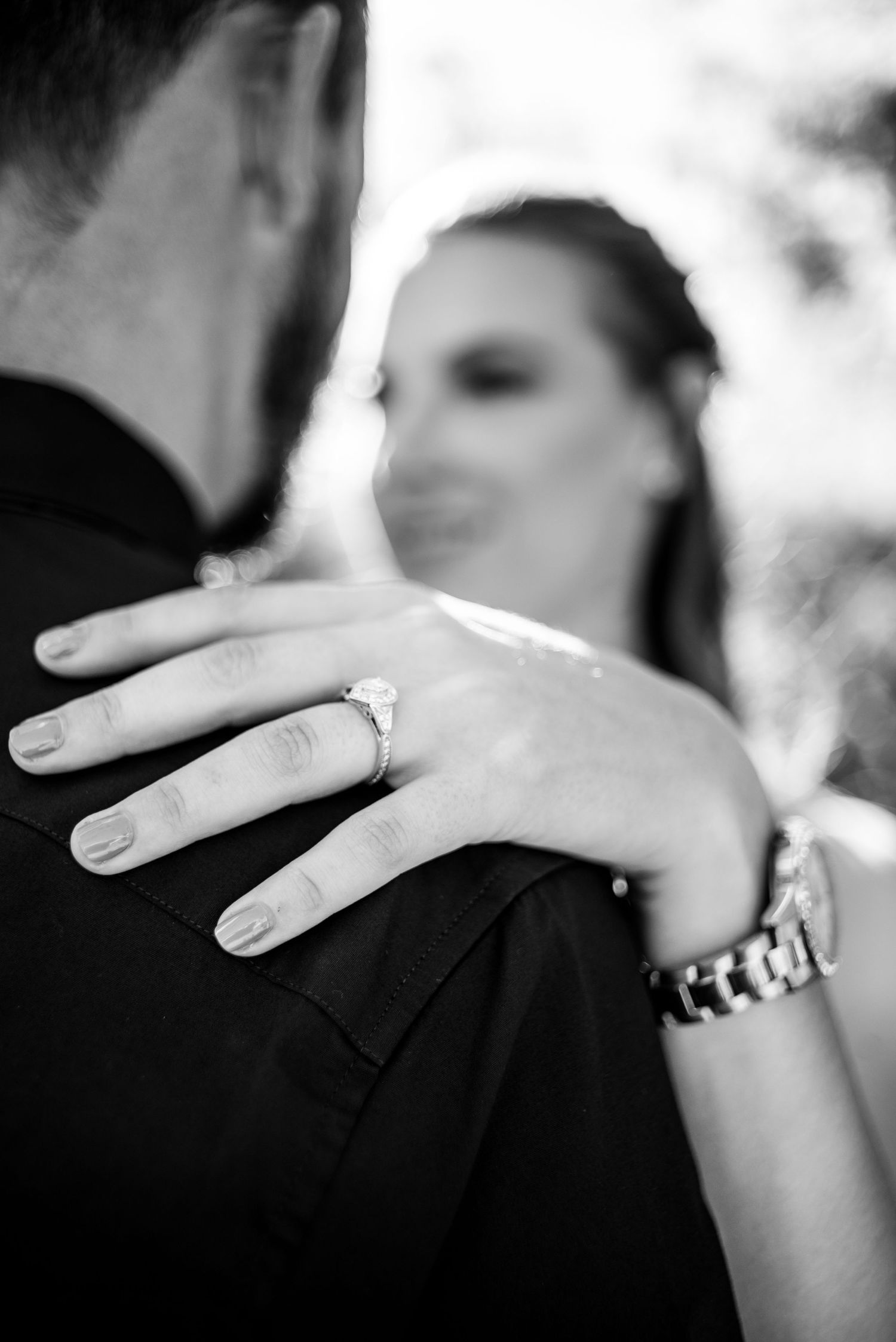 Black and white close-up of a bride's hand with engagement ring resting on groom's shoulder during wedding embrace.