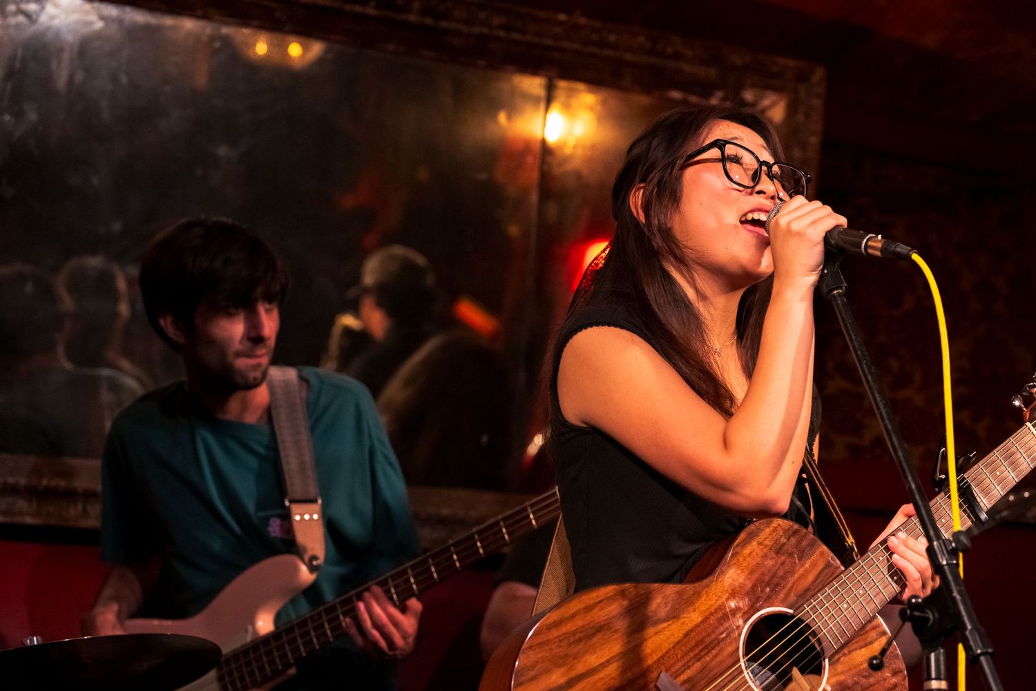Performers sing and play guitar at a dimly lit nightclub venue with warm red lighting and atmospheric mood.