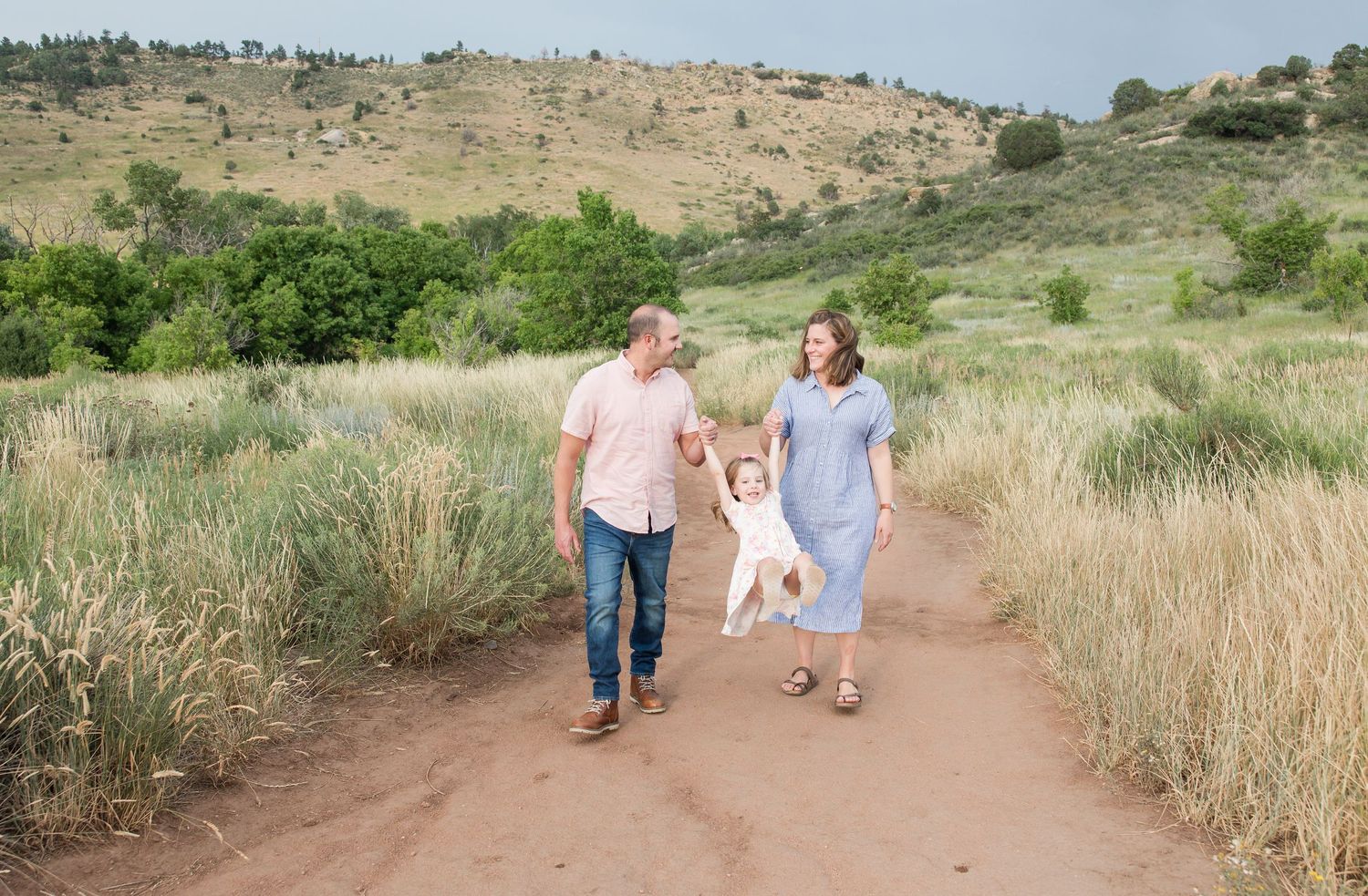 Family walking together on a dirt trail through golden grass with hills in the background during summer.