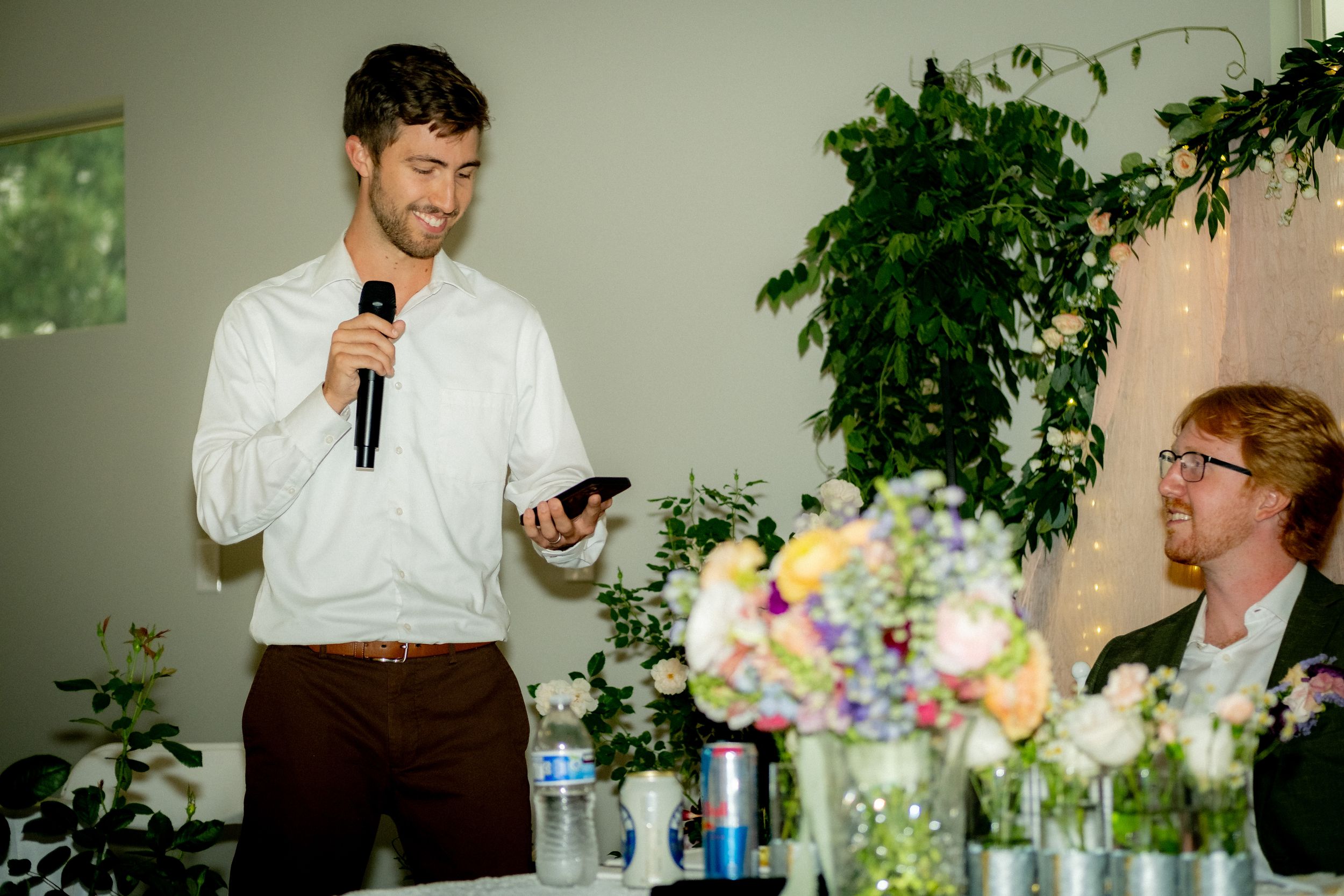 A speaker addresses wedding guests during a reception with floral centerpieces and greenery.