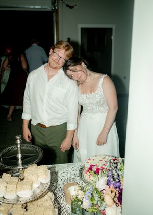 A couple stands beside their wedding dessert display with floral arrangements.