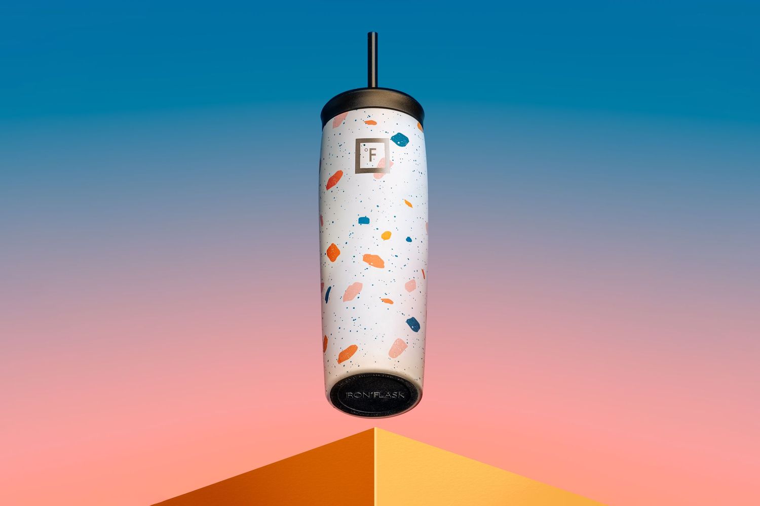 A white cylindrical lamp with colorful confetti pattern design floats against a gradient blue and coral background.