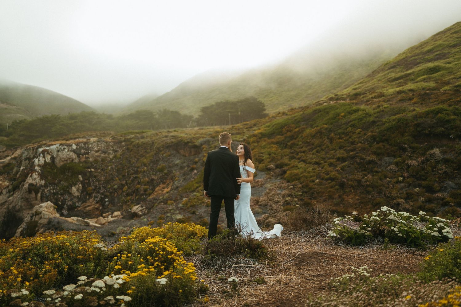 A romantic wedding couple stands alongside rolling coastal hills with foggy ocean views in the distance.