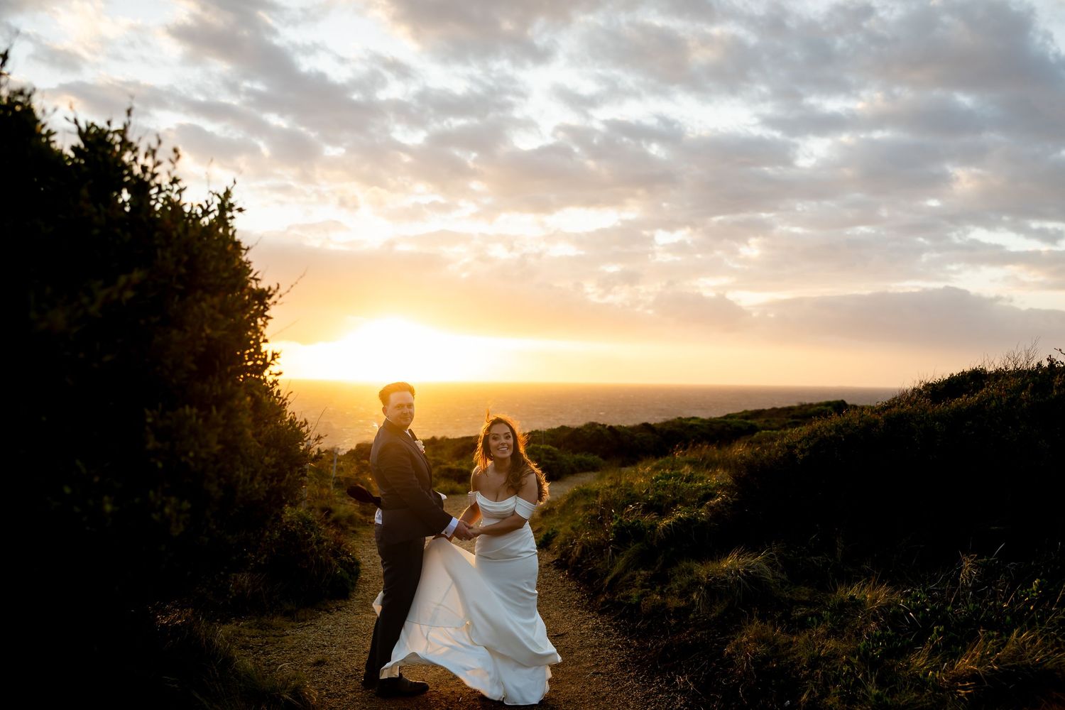 Bride and groom walk at golden sunset on coastal hillside, wedding dress flowing in breeze with ocean horizon backdrop.