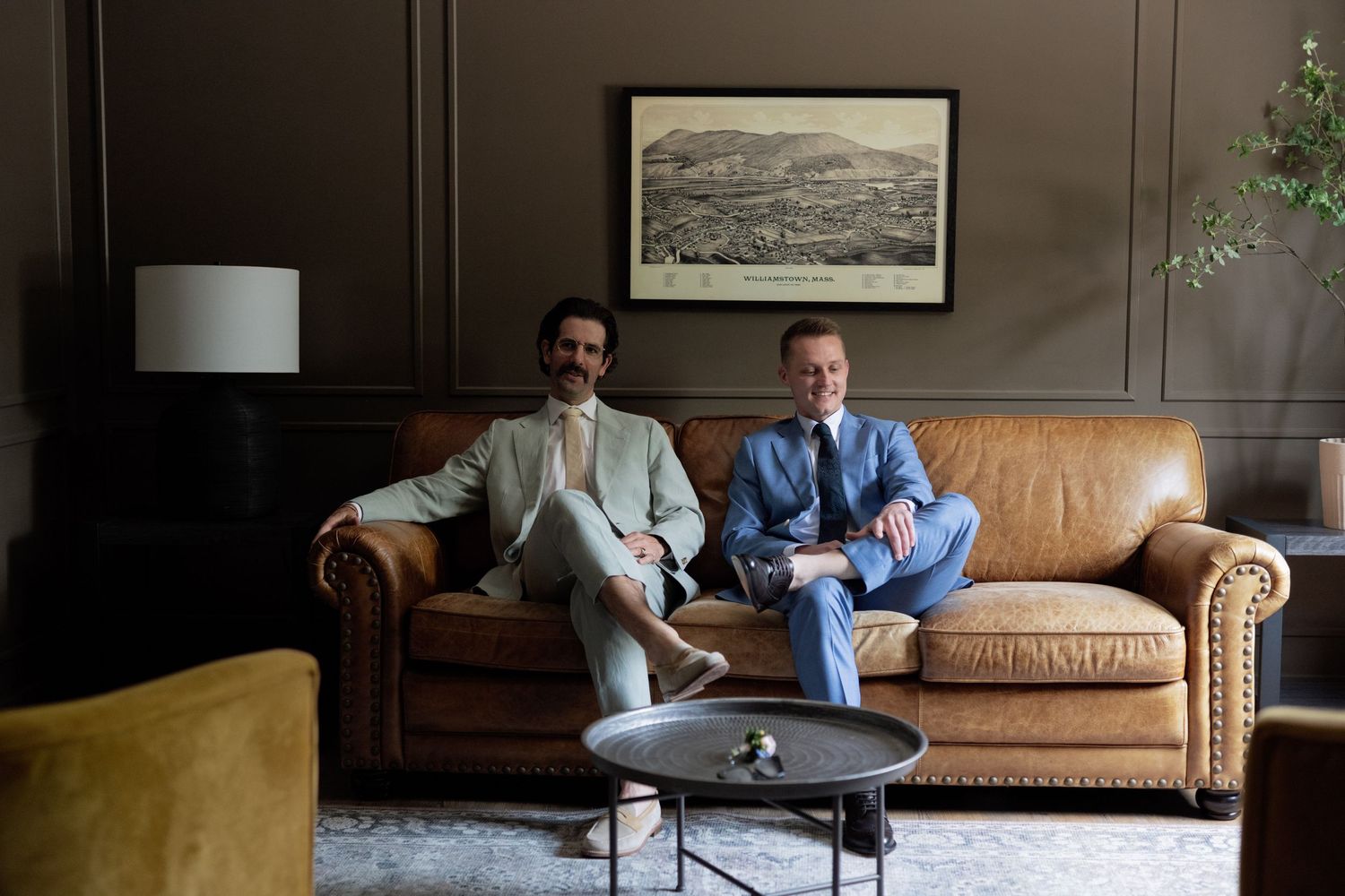 Two people sitting on a brown leather couch in a living room with artwork hanging on the wall behind them.