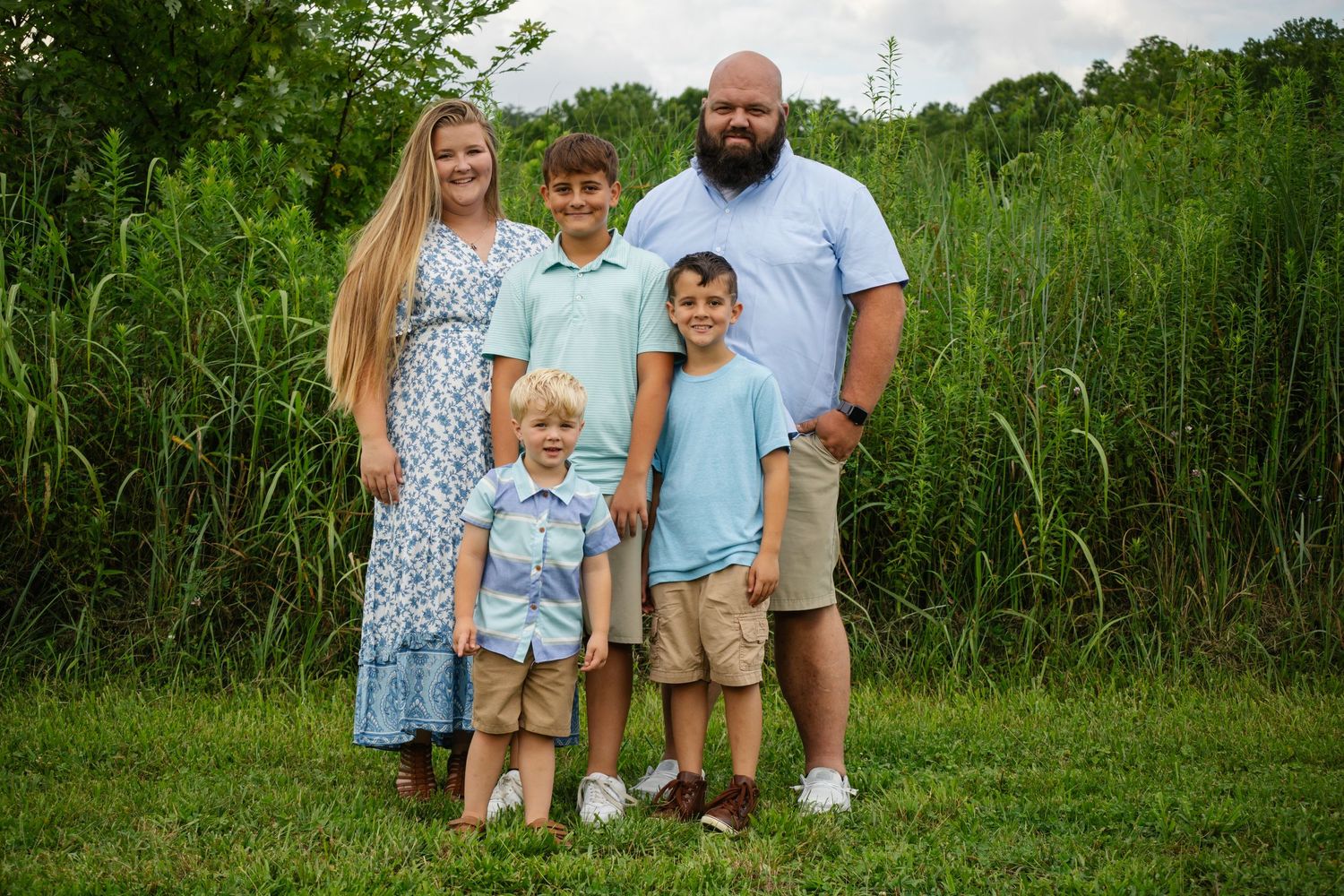 Family group poses together on grass with tall greenery and trees in background.