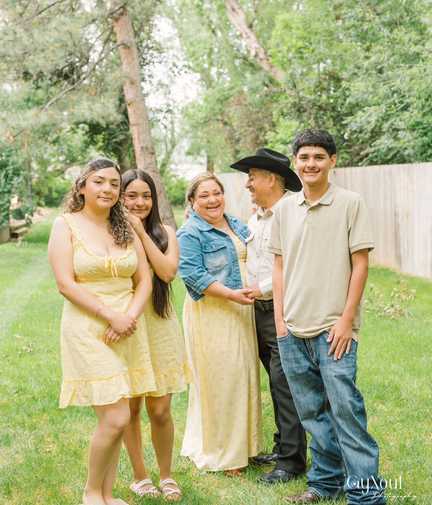 A family in casual attire gathers together for a portrait on a sunny day in their backyard garden.