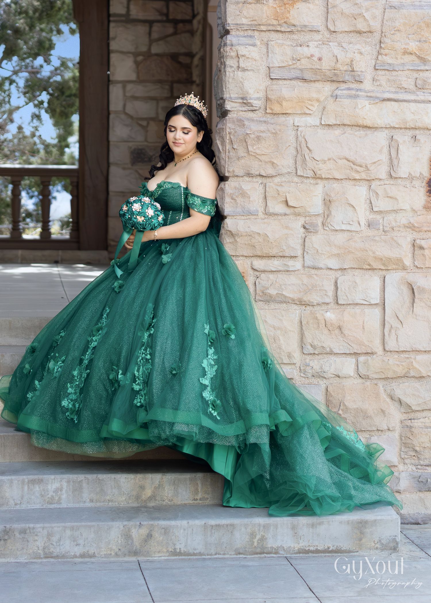 A stunning emerald green quinceañera ball gown with floral appliques is showcased against a stone wall backdrop.