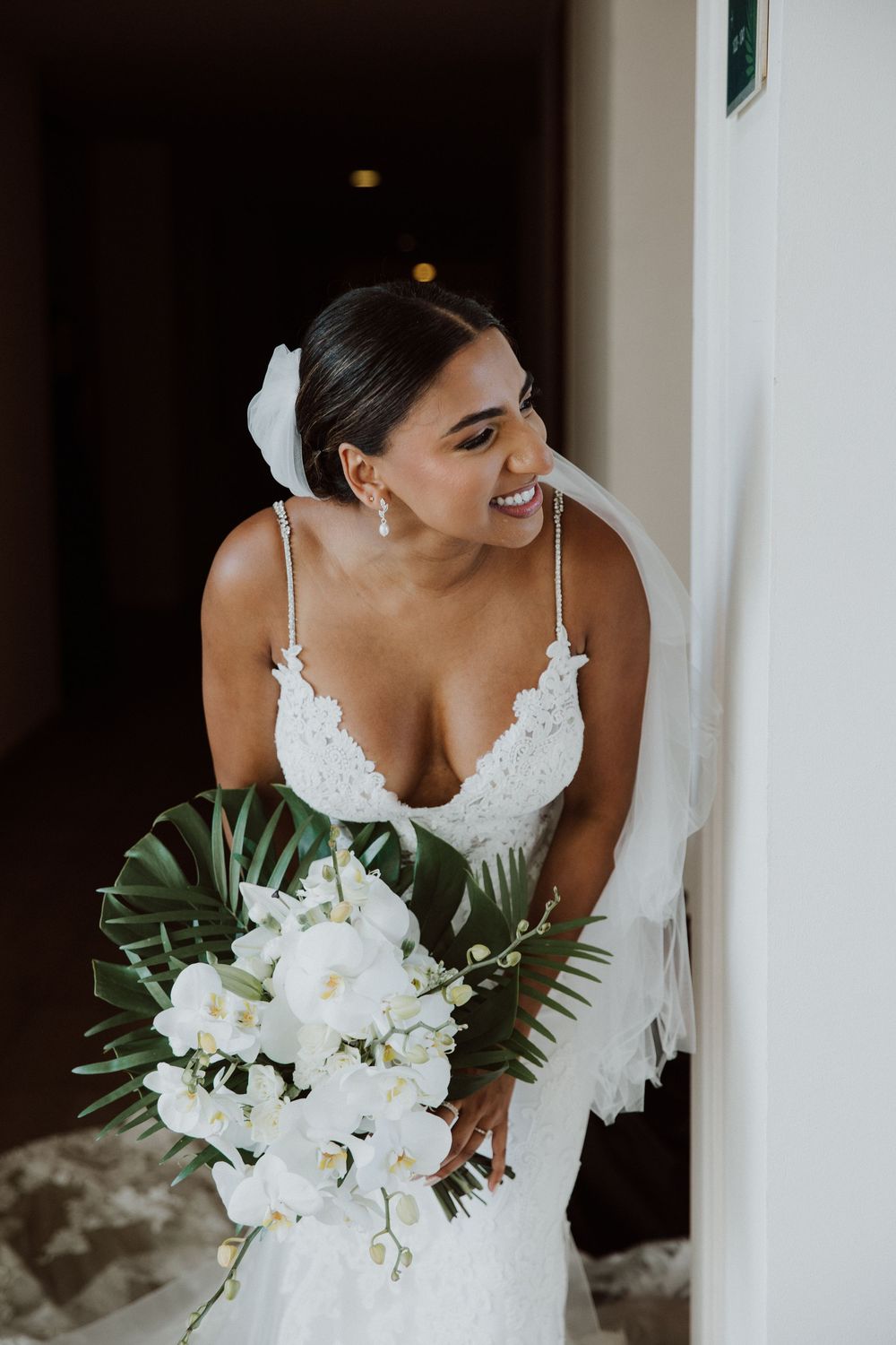 A bride in a white lace dress holds a tropical bouquet of white orchids and greenery.