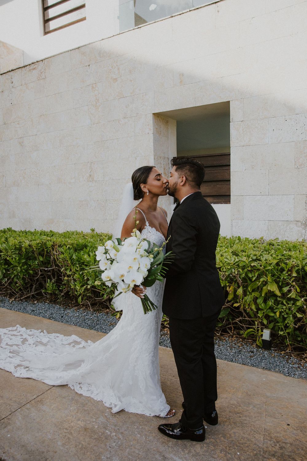 Romantic kiss between newlyweds against white stucco wall with tropical landscaping.