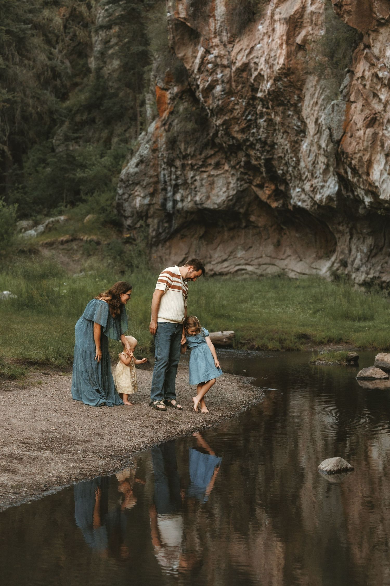 A family of three smiles together in a lush green field with a dramatic red rock cliff backdrop.