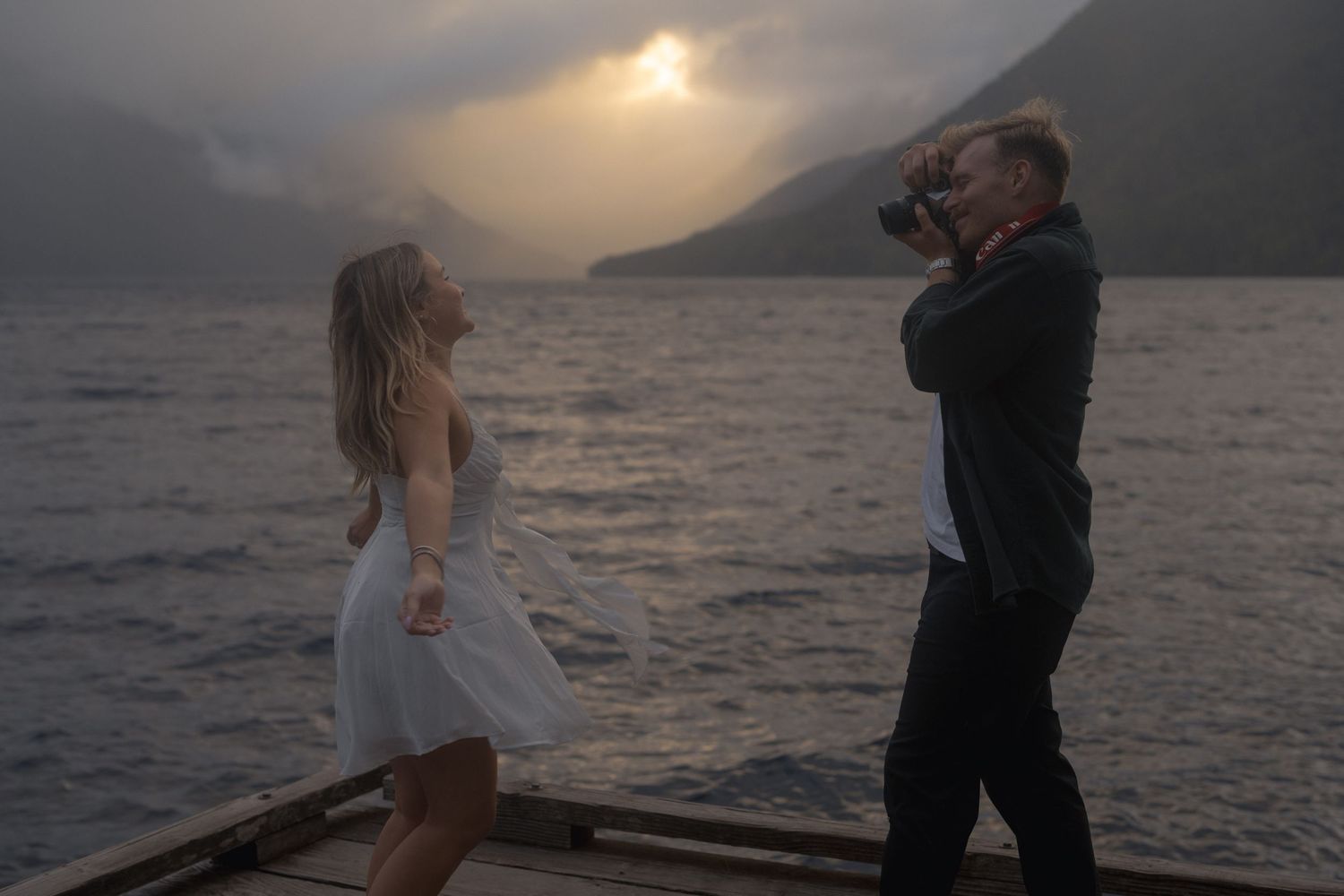 A photographer captures a scenic photoshoot on a boat deck at sunset with mountains and moody clouds over the ocean.