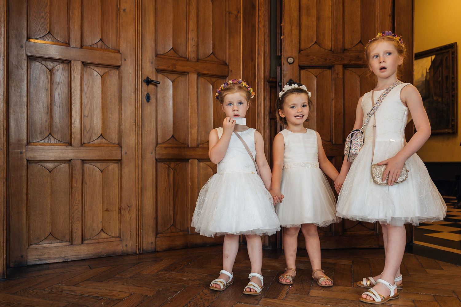Young flower girls in white tulle dresses sitting on wooden church bench.