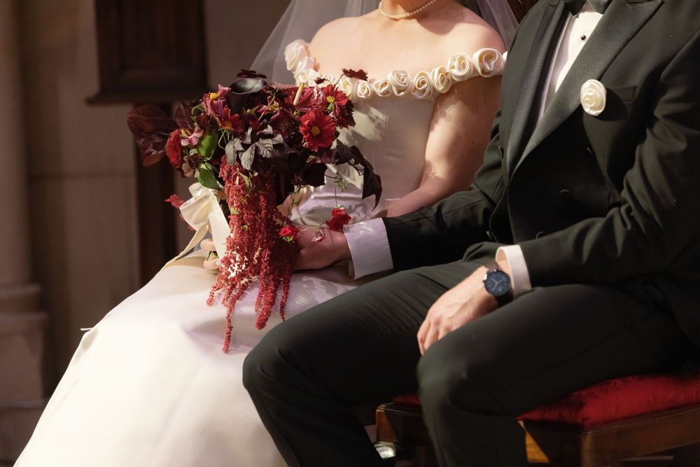 Elegant wedding scene showing a white ball gown dress and dark suit with a dramatic burgundy and red floral bouquet.