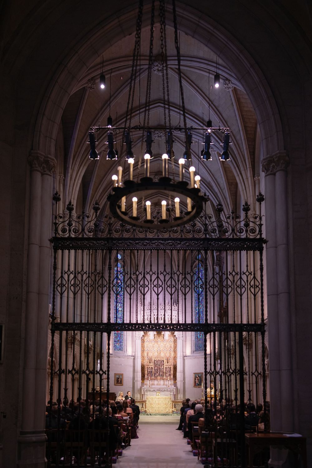 Gothic church interior with wrought iron gates and chandelier leading to illuminated altar.