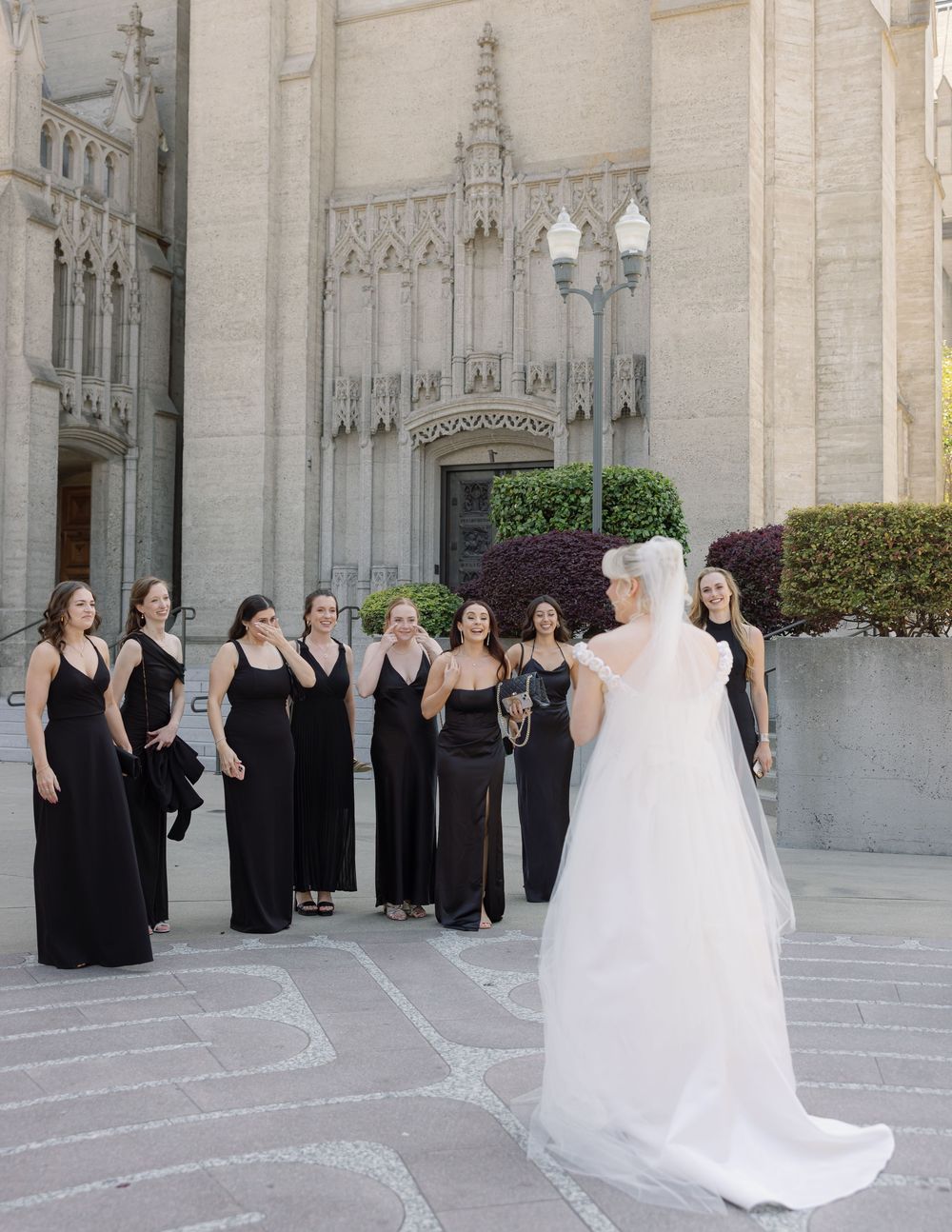 Wedding party in black formal dresses gathers outside a grand cathedral with ornate gothic architecture.