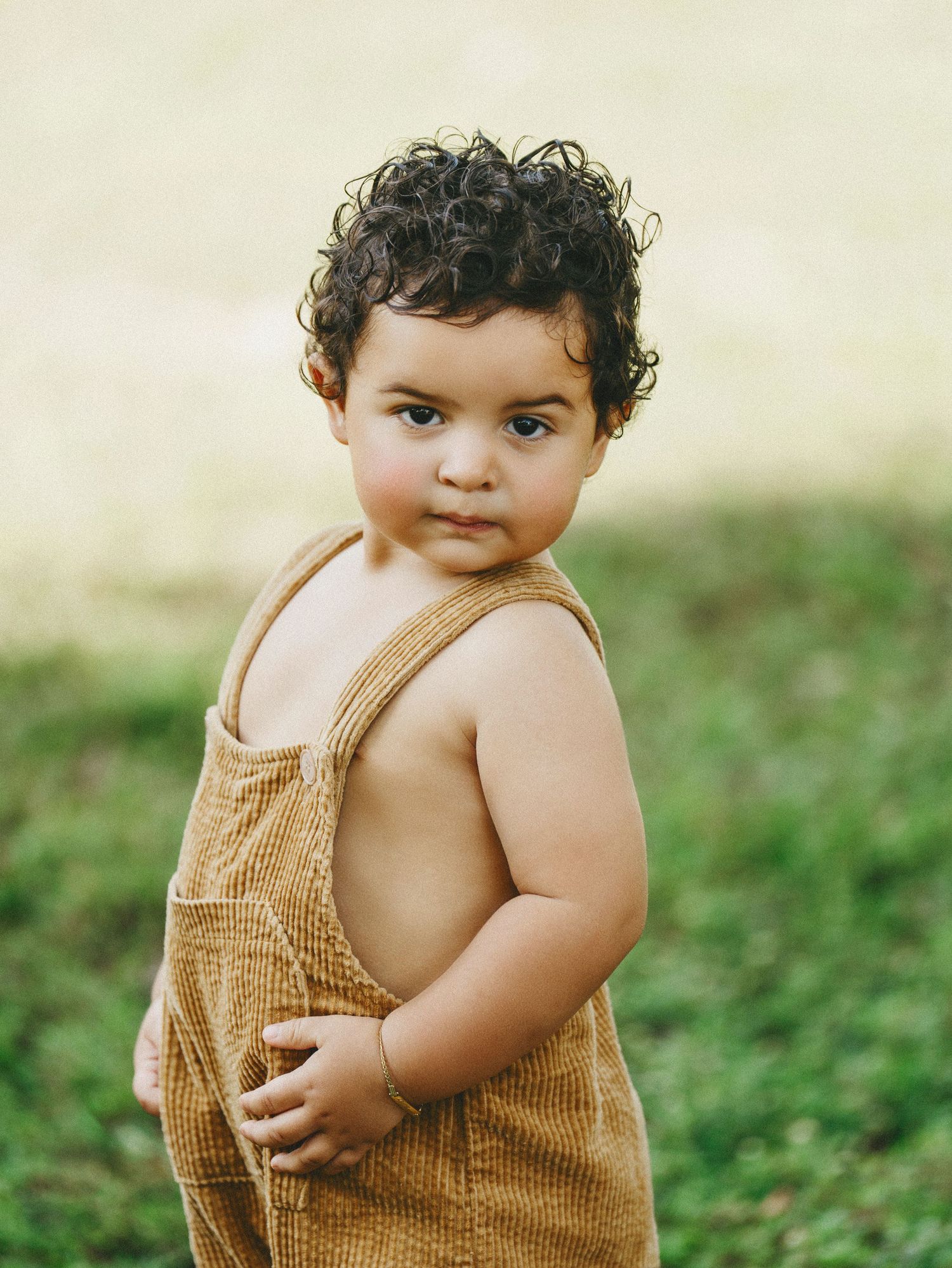 A toddler in tan overalls poses outdoors with curly hair and a sweet expression.