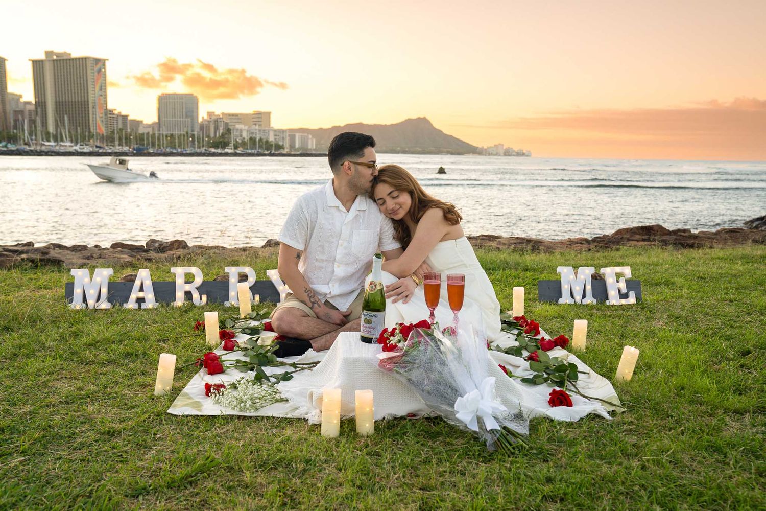 A romantic sunrise proposal picnic setup on grass at Magic island with "MARRY ME" letters in Ala Moana beach park, Honolulu.