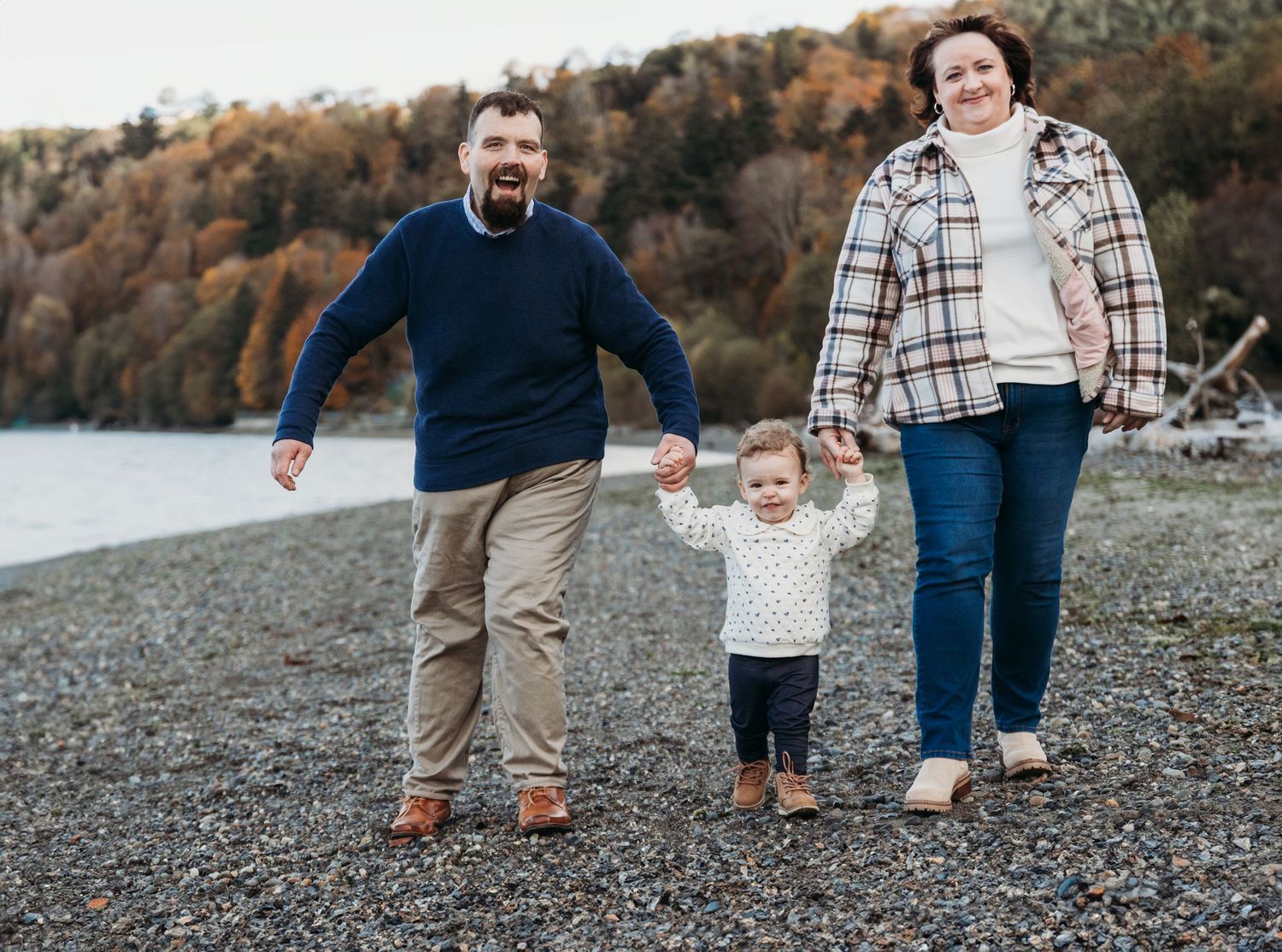 Three people walk together holding hands on a rocky beach shore during autumn with fall foliage in background.