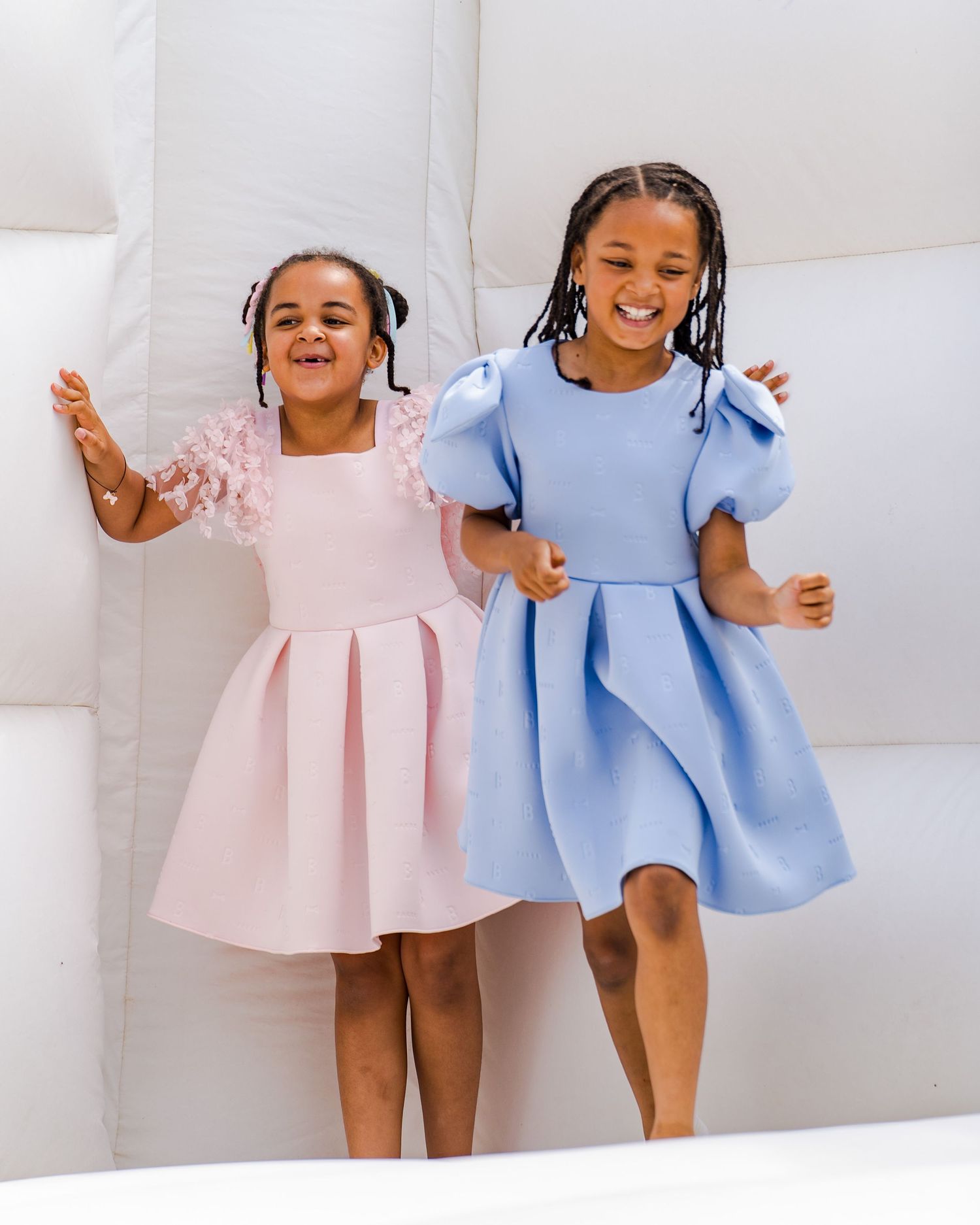 Two young children in pink and blue dresses pose playfully together on a white background.