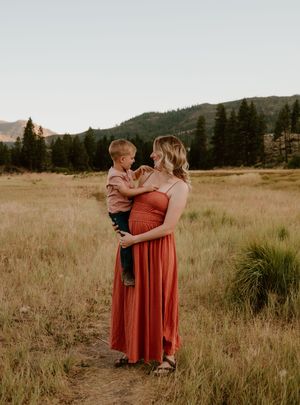 A figure in a rust-colored dress holds a small child while standing in a grassy meadow with mountains in the background.