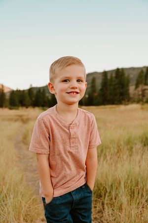 Young child playing in a grassy meadow wearing a pink t-shirt and dark pants against a mountain backdrop.