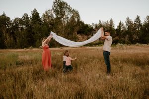 Three people stand in a golden field holding a flowing white veil as the sun sets behind pine trees.
