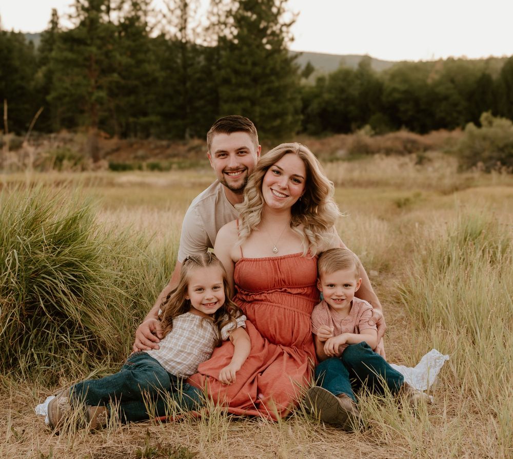 A loving family of four sits together in a golden grassy field during sunset, wearing coordinating casual outfits.