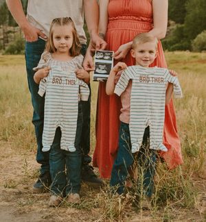 Two children holding up matching striped onesies in a field while adults hold an ultrasound photo in the background.