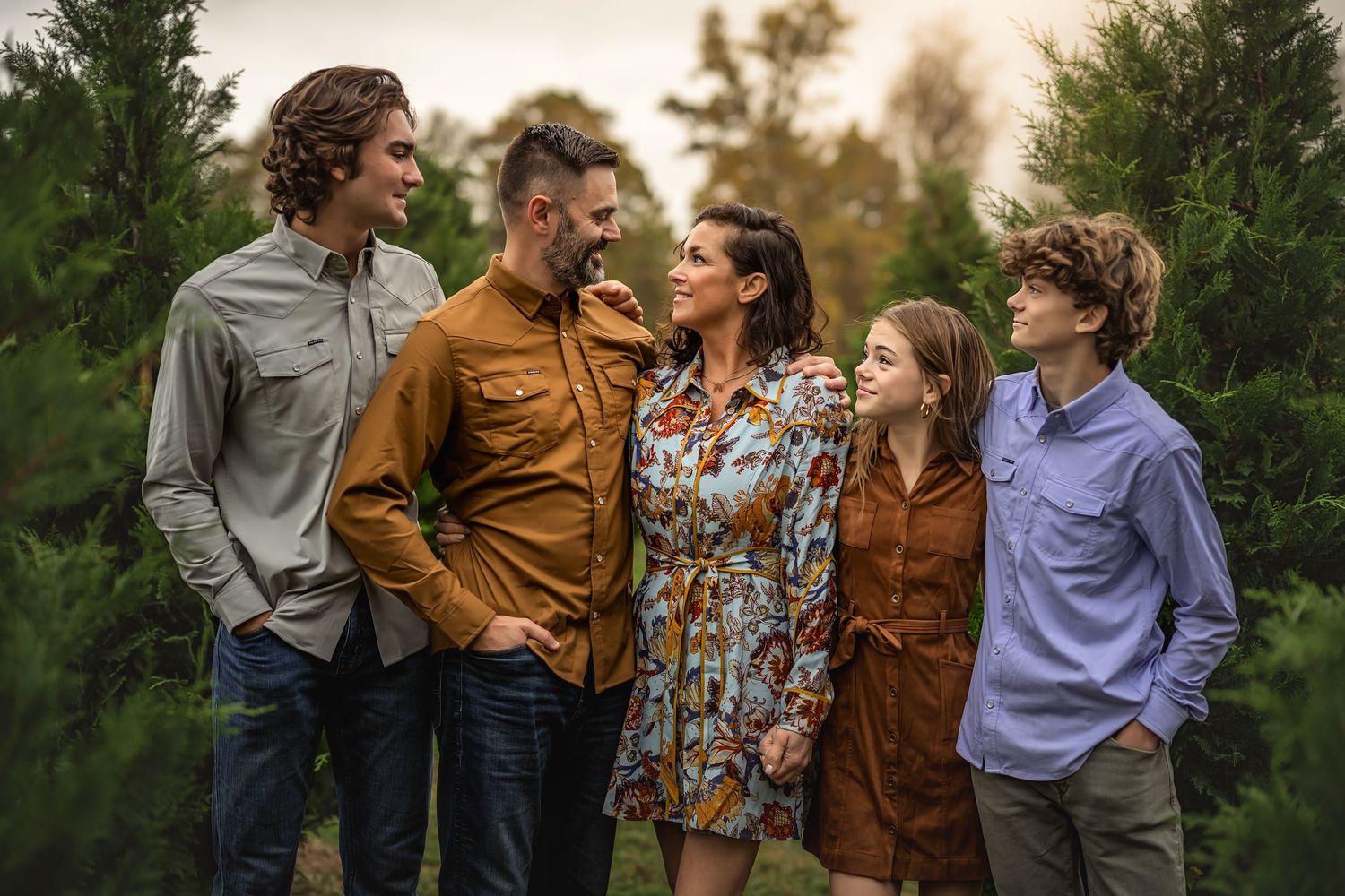 Family of five stand together outdoors against a backdrop of evergreen trees at sunset in casual fall attire.