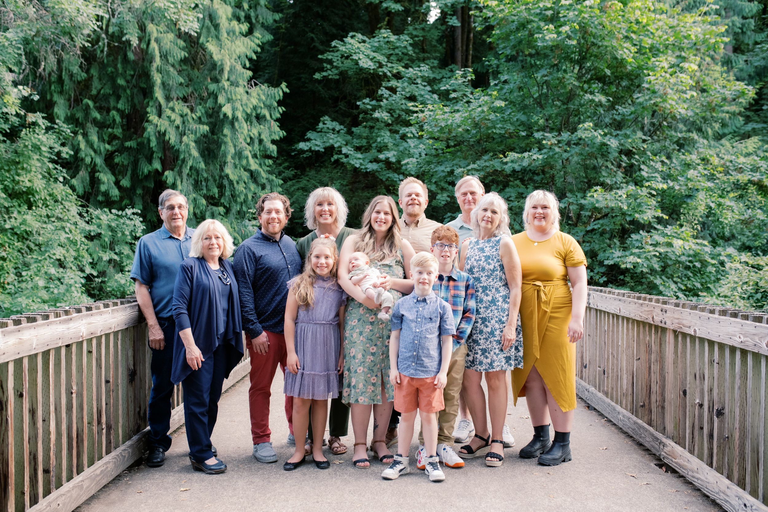 A group of people stand together on a wooden bridge walkway surrounded by lush green summer foliage.