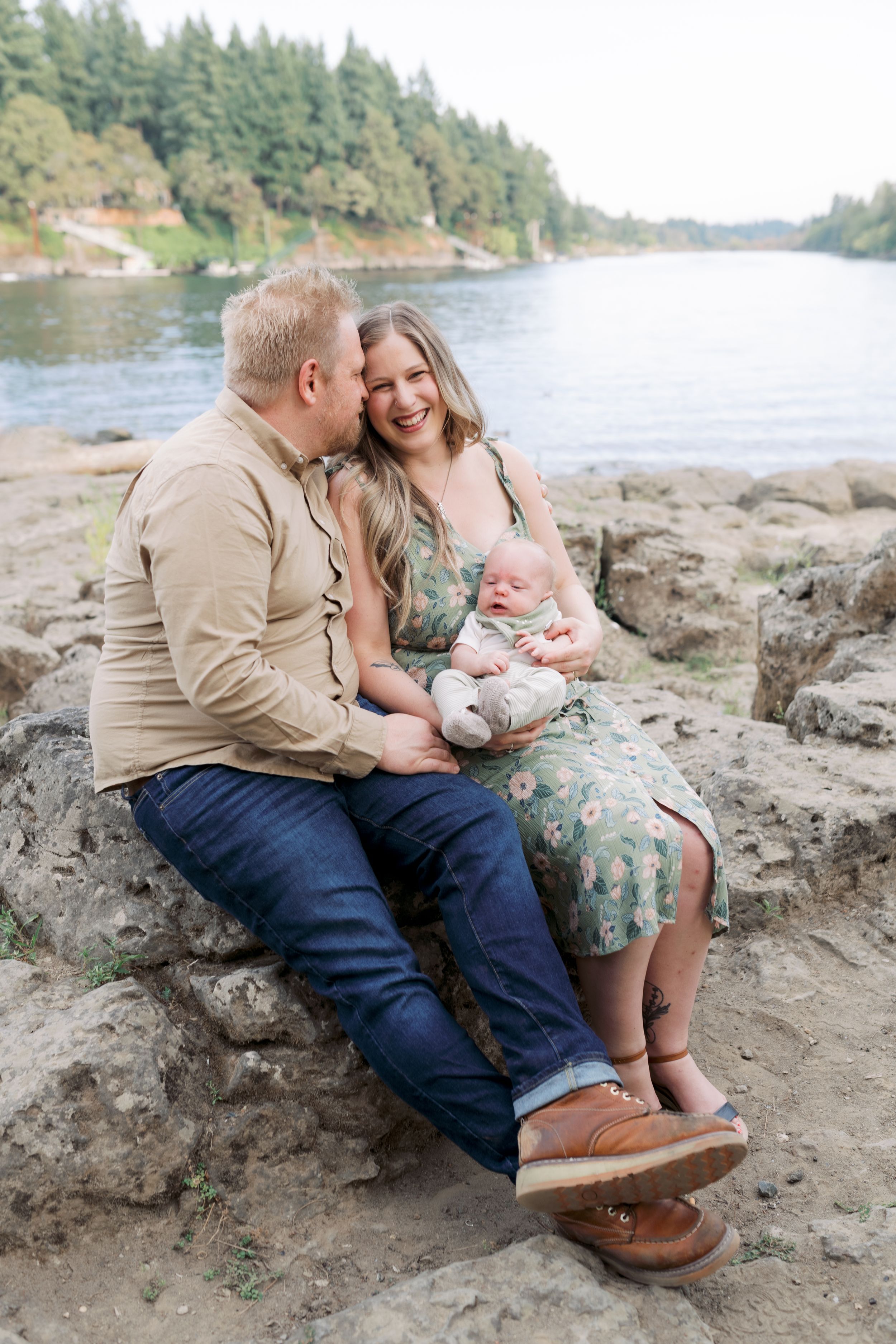 A romantic couple and infant sit together on rocky riverbank with evergreen forest and water in background.