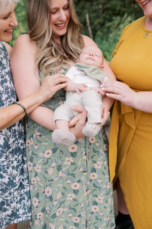 Three people dressed in floral and yellow dresses hold and smile at a baby wearing white clothing outdoors.