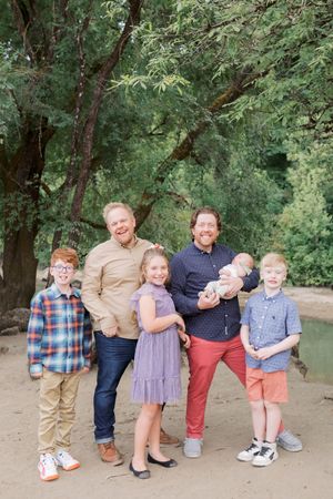 A family poses together on a dirt path surrounded by lush green trees in an outdoor setting.