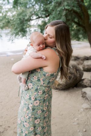 A parent wearing a floral dress holds and kisses a baby on a sunny beach under trees.
