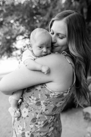 A tender black and white portrait shows a mother in a floral dress embracing and kissing her baby outdoors.